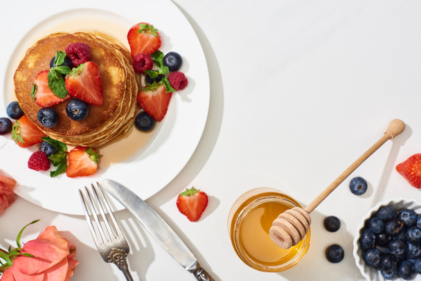 felülnézet ízletes palacsinta mézzel, áfonyával és eper tányéron közel evőeszközök és rózsa márvány fehér felületen - Fotó, kép