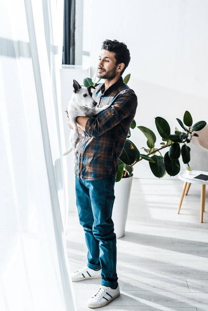 handsome bi-racial man holding Jack Russell Terrier and looking through window  - Fotó, kép