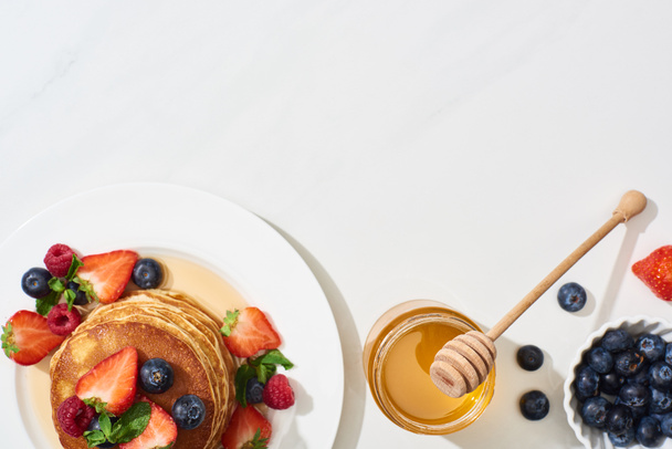 vista superior de deliciosas panquecas com mel, mirtilos e morangos na placa na superfície branca de mármore
 - Foto, Imagem