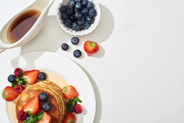 top view of delicious pancakes with blueberries and strawberries on plate near maple syrup in gravy boat on marble white surface - Photo, Image