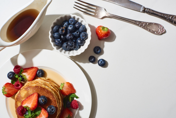 vue de dessus de délicieuses crêpes aux bleuets et fraises sur assiette près des couverts et du sirop d'érable en saucière sur surface marbre blanc
 - Photo, image