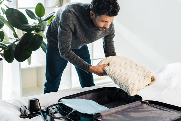 handsome bi-racial man in sweater packing travel bag in apartment  - Photo, Image