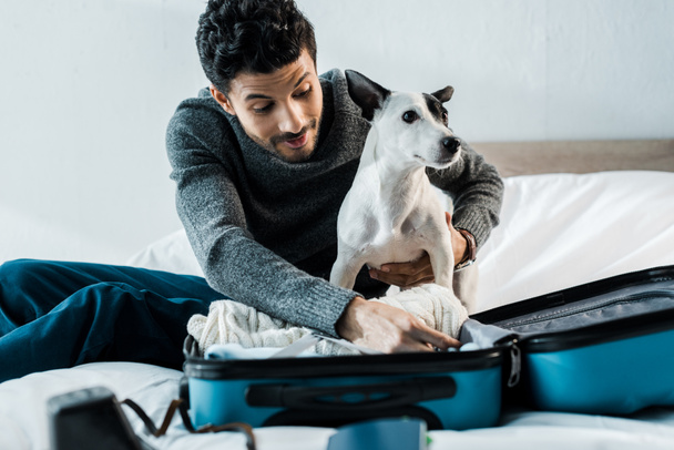 handsome and smiling bi-racial man packing luggage and holding jack russell terrier - Φωτογραφία, εικόνα