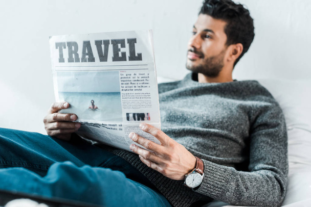 selective focus of handsome and smiling bi-racial man holding newspaper with lettering travel  - Foto, Imagen