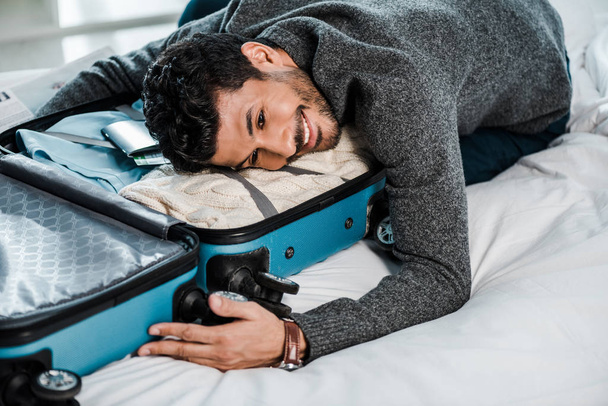 Yakışıklı ve gülümseyen çift ırklı bir adam seyahat çantasında yatıyor. - Fotoğraf, Görsel
