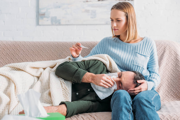 attentive wife looking at thermometer near diseased husband sneezing while lying on her laps - Photo, Image