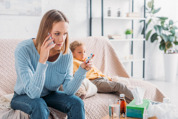 madre preocupada sosteniendo ampolla de píldoras y hablando en el teléfono inteligente mientras está sentado cerca de hijo enfermo
 - Foto, Imagen