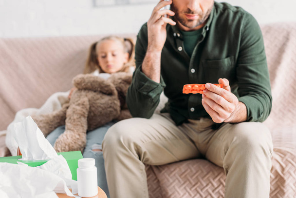 visão cortada do homem falando no smartphone e segurando bolha de pílulas enquanto sentado perto da filha doente
 - Foto, Imagem