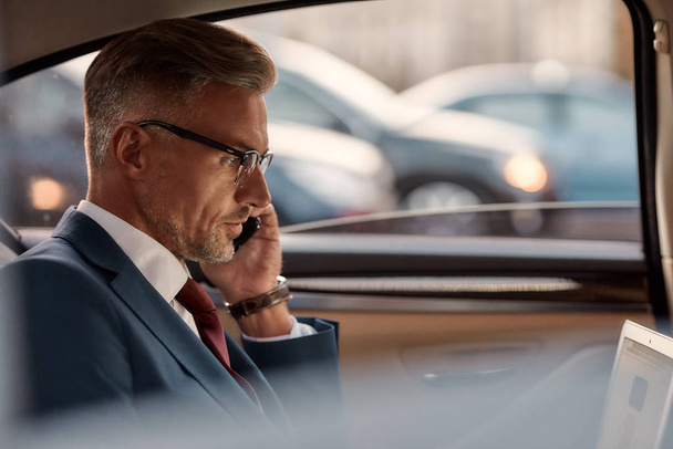 Busy businessman. Side view of mature man in classic wear sitting in the car and talking by the phone with client - Foto, immagini