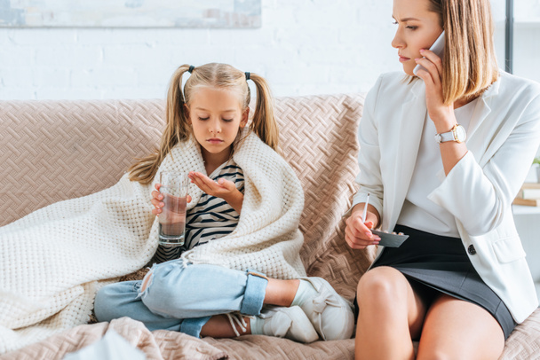sick child holding glass of water and pills near mother talking on smartphone - Foto, afbeelding