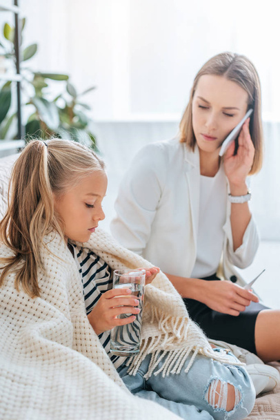 sick child holding glass of water and pills near mother talking on smartphone - Foto, Bild