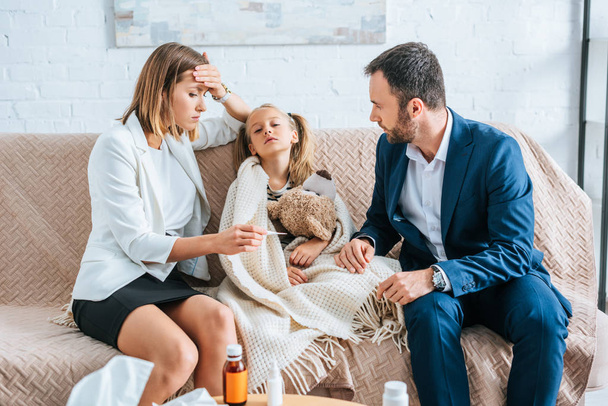 worried mother touching forehead and looking at thermometer while sitting near diseased daughter and husband - Foto, afbeelding