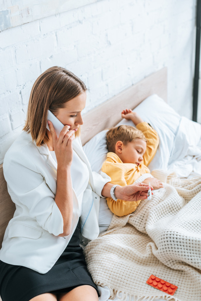 huolestunut äiti katselee lämpömittaria ja puhuu älypuhelimella istuessaan lähellä sairasta poikaa
 - Valokuva, kuva