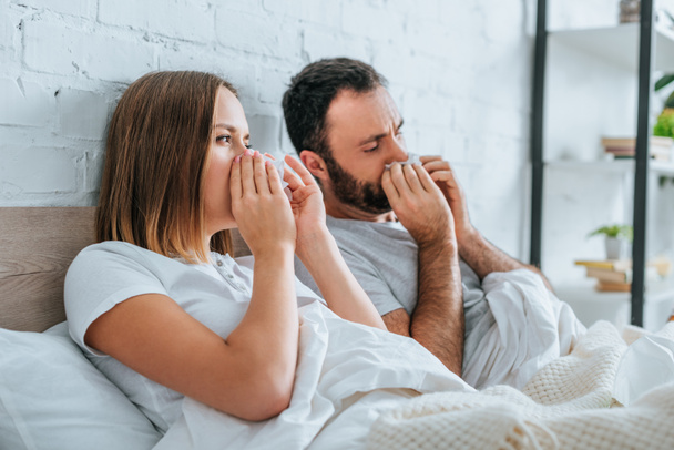 sick husband and wife sneezing while lying in bed together - Photo, Image