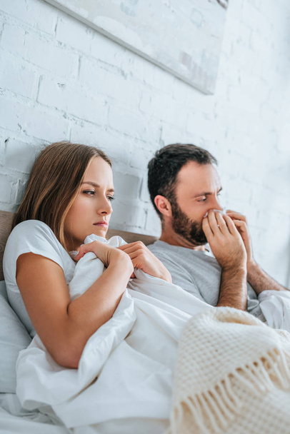 Erkrankter Mann niest in Serviette, während er neben kranker Frau im Bett liegt - Foto, Bild