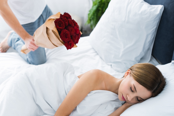 καλλιεργημένη άποψη του άνδρα κρατώντας μπουκέτο και γυναίκα κοιμάται στο κρεβάτι  - Φωτογραφία, εικόνα