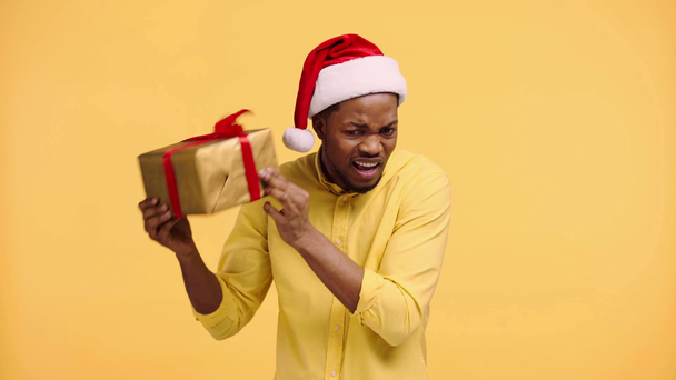 nieuwsgierig Afrikaans amerikaanse man schudden geschenk doos geïsoleerd op geel - Video