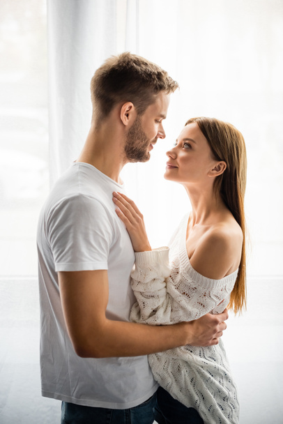 side view of handsome man hugging attractive and smiling woman in apartment  - Fotografie, Obrázek