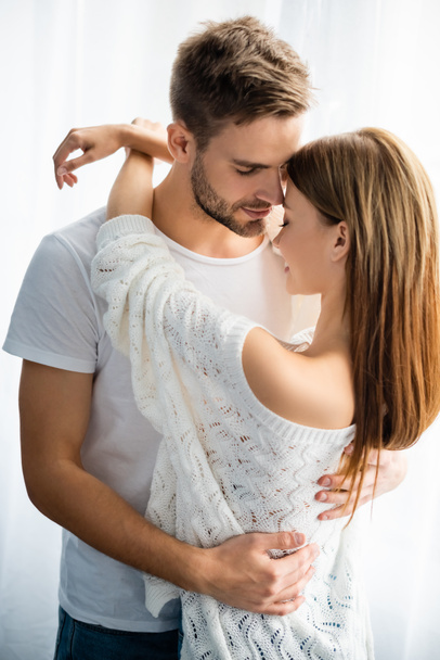 handsome and smiling man hugging woman in apartment  - Fotografie, Obrázek