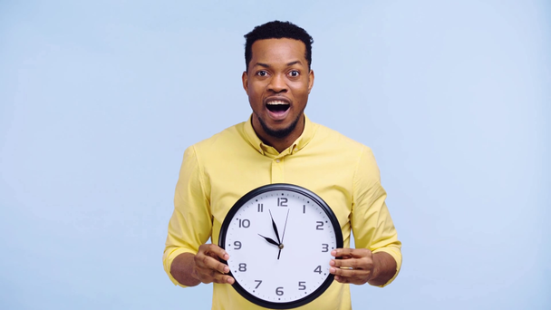 surprised african american man holding clock isolated on blue - Кадри, відео