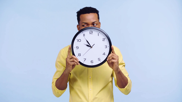 cheerful african american man holding clock isolated on blue - Кадри, відео