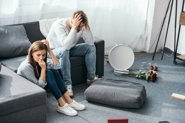 sad woman sitting on floor and handsome man sitting on sofa in robbed apartment  - Zdjęcie, obraz