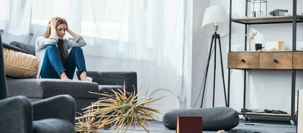 panoramic shot of attractive and sad woman sitting on sofa in robbed apartment  - Foto, imagen