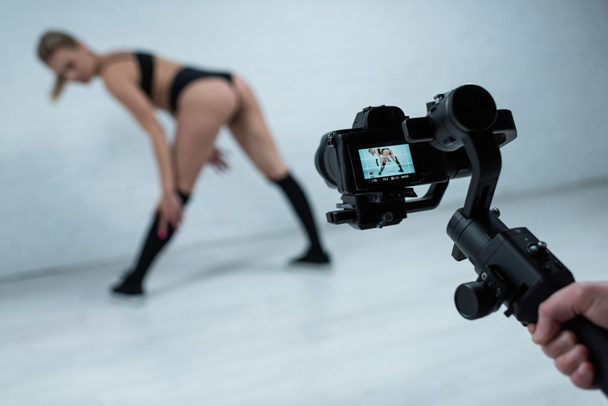 foco seletivo de cameraman tiro menina loira sexy em roupa preta twerking perto da parede de tijolo branco
 - Foto, Imagem