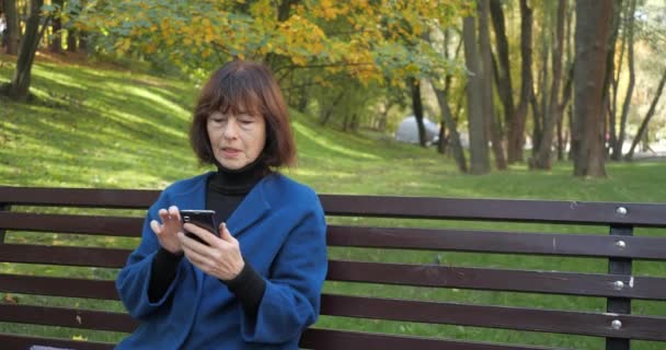 Elderly woman typing a phone message while sitting at the autumn park. Happy old woman looking at screen on smartphone. Senior woman using wireless internet connection on smart phone in slow motion. - Filmagem, Vídeo