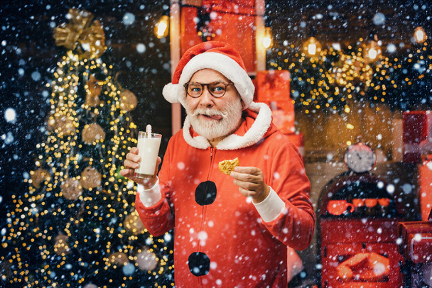 Porträt eines bärtigen komischen Mannes im Weihnachtsmannkostüm. Weihnachtsmann Claus genießt Plätzchen und Milch, die ihm an Heiligabend weggelassen werden. Milch und Kekse für den Weihnachtsmann. Weihnachtsmann pflückt Plätzchen. - Foto, Bild