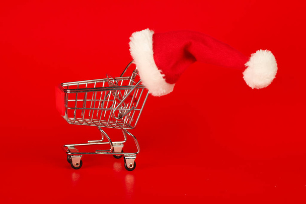 Shopping cart with Christmas Santa hat on red background. Xmas s - Foto, imagen