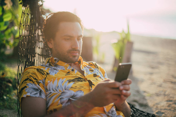 Молодой человек лежит в гамаке на солнечном пляже с телефоном
. - Фото, изображение