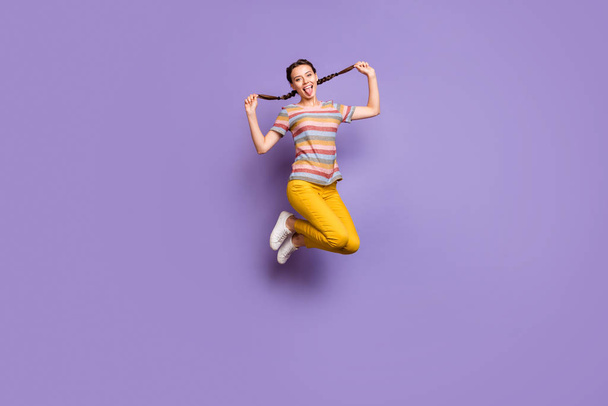 Full length photo of funny lady jumping high summer mood holding pretty braids sticking tongue out mouth wear casual striped t-shirt yellow pants isolated purple color background - Photo, Image