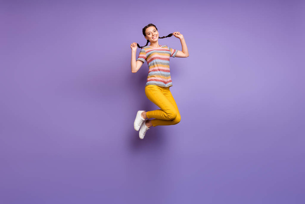 Full length photo of crazy lady jumping high in amazing summer mood holding pretty braids wear casual striped t-shirt yellow pants isolated purple color background - Foto, Imagen