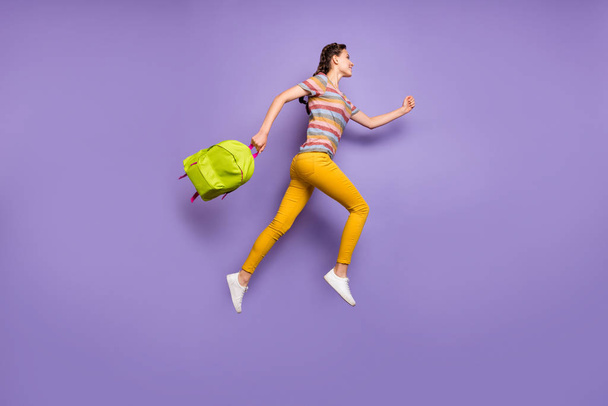 Full size photo of crazy excited lady jumping high carry school green rucksack rushing home after lessons wear casual striped t-shirt yellow pants isolated purple color background - Фото, изображение