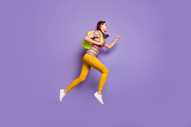 Full length profile photo of funky lady jumping high carry school bag rushing home after classes wear casual striped t-shirt yellow trousers isolated purple color background - Фото, зображення