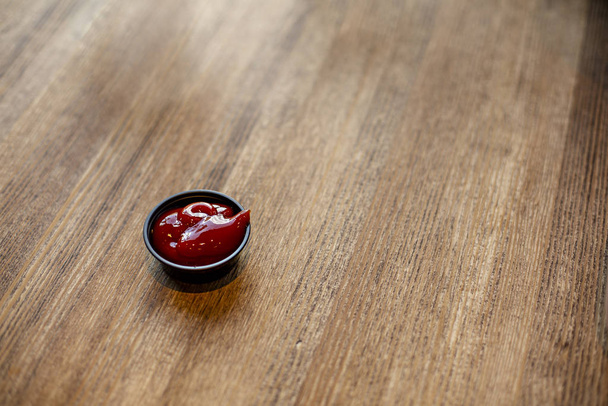 Cierre de un pequeño contenedor de Ketchup sobre una mesa de madera. Vista de enfoque selectiva sobre la salsa de ketchup en el recipiente. - Foto, imagen