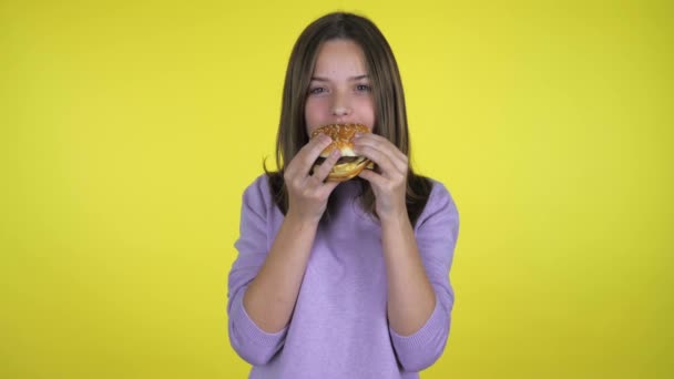 Dospívající dívka v růžovém svetru jí burger a dívá se do kamery. Žluté pozadí s kopírovacím prostorem. Nezdravé jídlo. Zdravý stravovací koncept. Emoce teenagerů. Snímek zpomalení 4k - Záběry, video