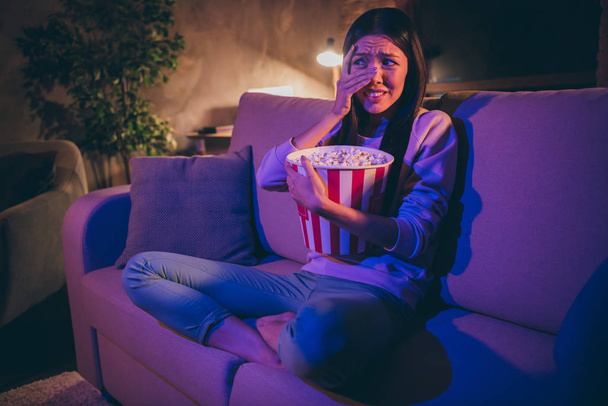 Photo of homey chinese lady eat popcorn movie night watch horror scary movie hiding eyes afraid look on blood sit cosy couch casual outfit dark living room indoors - 写真・画像
