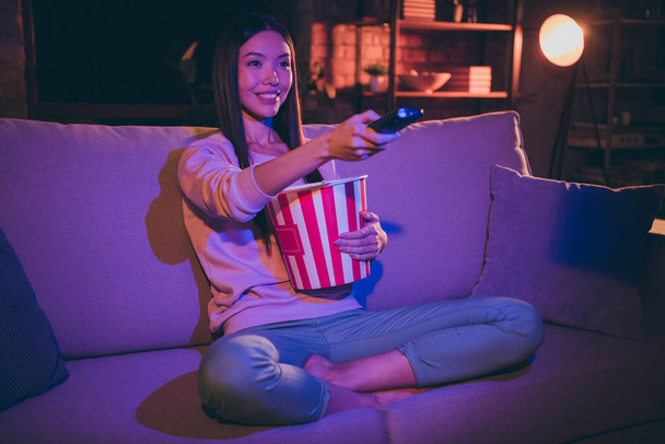 Photo of pretty lady domestic mood holding tv remote controller changing channels eating popcorn movie night sitting comfy couch casual outfit dark living room indoors - Foto, immagini