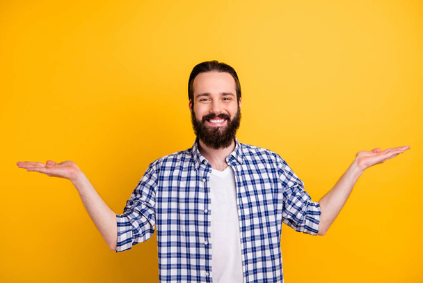 Portrait of his he nice attractive cheerful cheery bearded guy in checked shirt holding on palms two invisible products deciding isolated on bright vivid shine vibrant yellow color background - 写真・画像