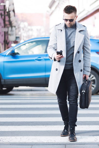 Moderně oblečený obchodník spěchá přes ulici, dívá se na mobil a nese notebook. Koncept dnešního rušného života plného technologií - Fotografie, Obrázek