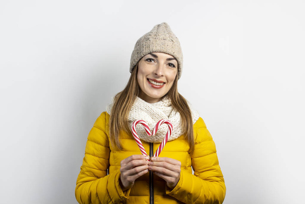 Young woman in a hat and a down jacket holds a candy caramel cane on a light background. Heart shape. Concept winter, christmas, winter holidays - Foto, afbeelding