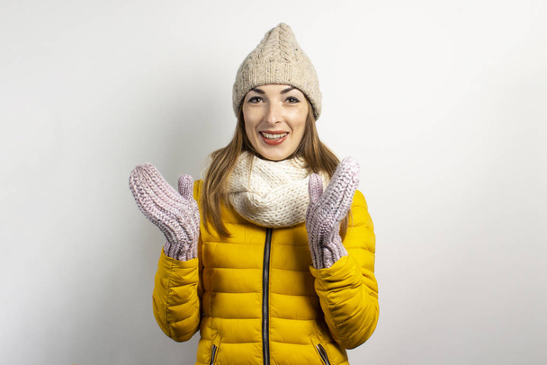 Jovem mulher em um chapéu quente e luvas está sorrindo em um fundo leve. Conceito de inverno, moda de inverno, roupas de inverno. Humor engraçado, emoções, surpresa, choque - Foto, Imagem