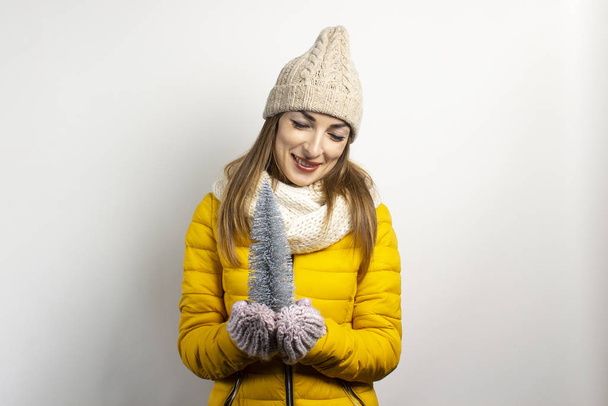 Young woman in a warm hat and mittens smiles and holds a decorative Christmas tree on a light background. Concept of winter, winter holidays, Christmas, New Year. Funny mood, emotions, surprise - Photo, Image