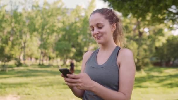 Caucasian fit young woman smiling while texting on mobile phone at the park in the morning on a sunny day - Metraje, vídeo
