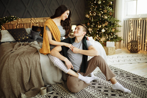 happy couple in love in the morning on the bed near the Christmas tree, hug, Christmas morning. home clothes and sweaters. comfort and warmth of hearts - Photo, Image