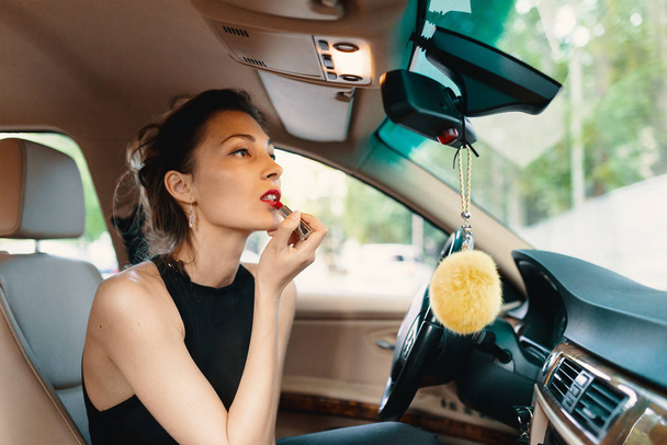 Молода елегантна жінка дивиться в дзеркало перегляду автомобіля під час нанесення макіяжу, помади на губи
 - Фото, зображення