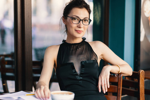 Молода стильна жінка зі стильними окулярами сидить за столом у кафе з великою чашкою кави або латте
 - Фото, зображення