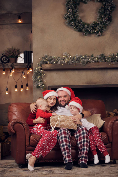 Image of young parents in Santas caps and sons on red leather sofa in studio - Foto, Imagem